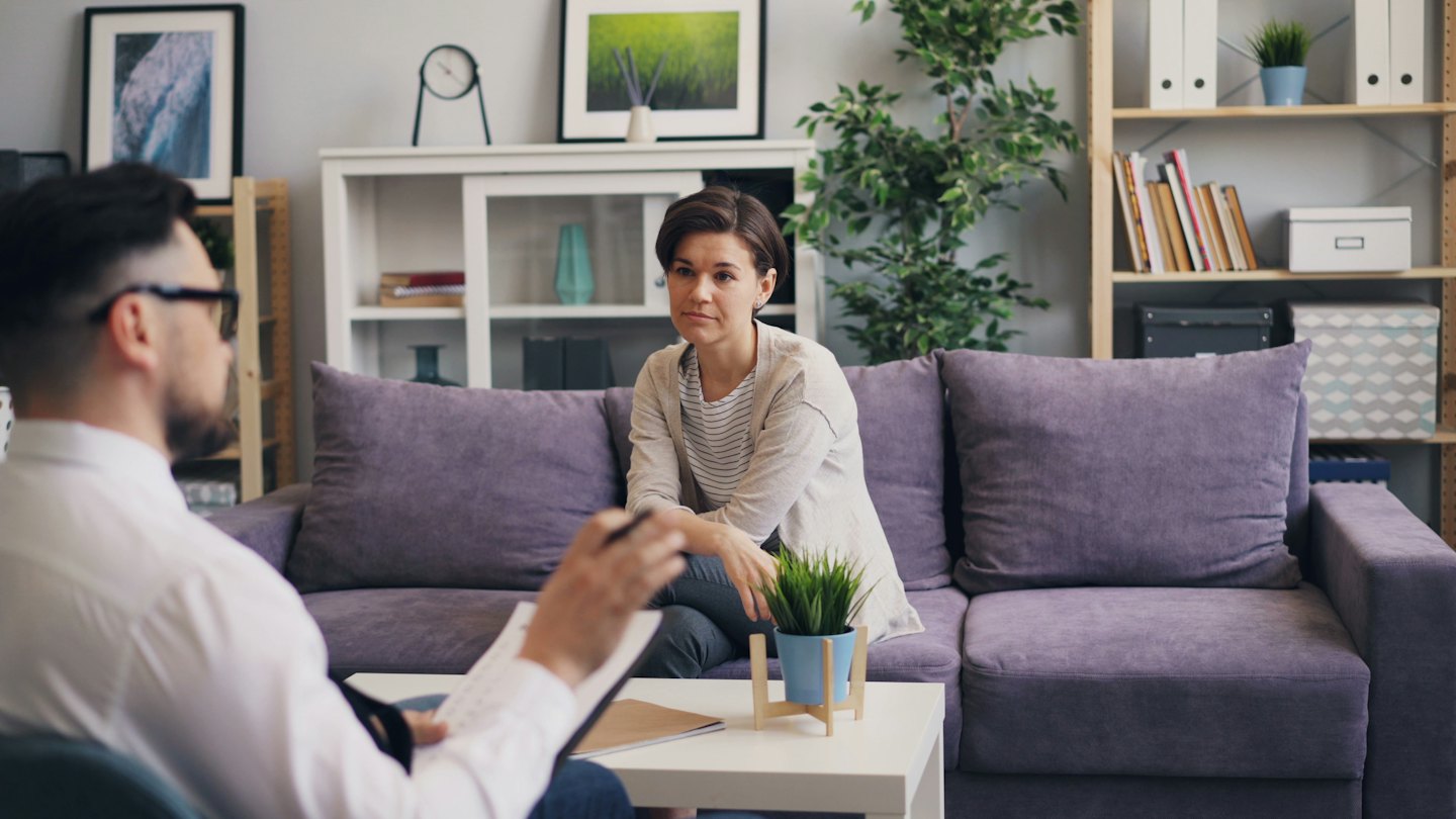 Therapist on clipboard listens to woman on purple sofa.