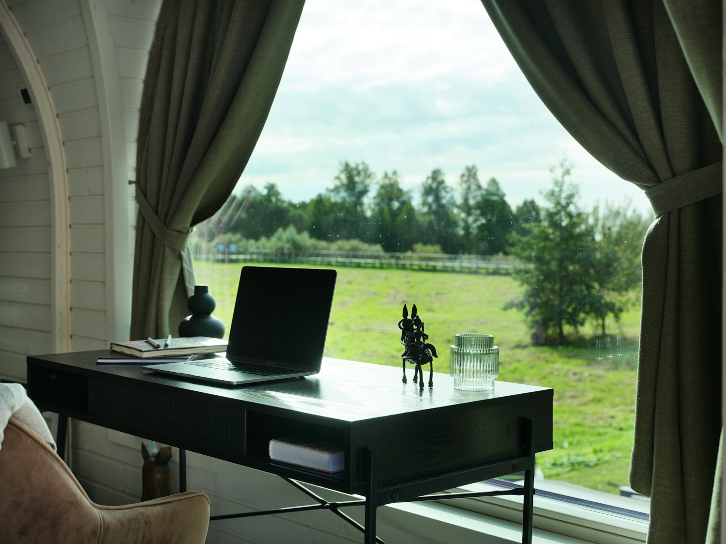 Laptop on a wooden desk next to a window with a green field view.